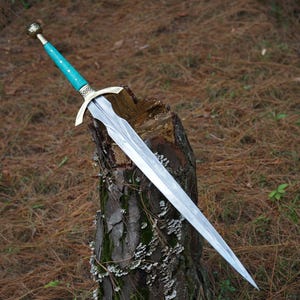 May include: A silver sword with a teal handle and gold accents, resting on a tree stump. The blade is double-edged and tapers to a point. The hilt features a crossguard and a decorative pommel.