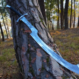 Puede incluir: Una espada plateada con mango negro se apoya en un tronco de &aacute;rbol en un entorno boscoso.