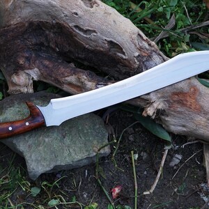 May include: A long, silver-colored sword with a wooden handle. The handle is brown with a dark wood grain and has a slight curve. The sword is lying on a piece of wood and some green grass.