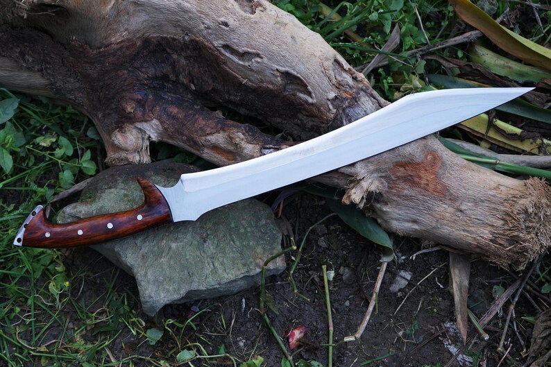 May include: A long, silver-colored sword with a wooden handle. The handle is brown with a dark wood grain and has a slight curve. The sword is lying on a piece of wood and some green grass.