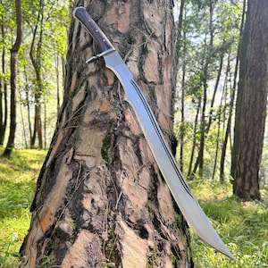 Könnte beinhalten: Ein großes, gebogenes Schwert mit polierter silberner Klinge und dunklem Holzgriff. Die Klinge hat drei parallele schwarze Linien. Das Schwert lehnt an einem Baum in einer Waldumgebung.