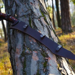 May include: A brown leather scabbard with a long, curved blade. The scabbard has a flap with a snap closure and is secured to a tree trunk.