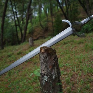 May include: A silver-toned metal sword with a double-edged blade, a crossguard with curved quillons, and a textured handle featuring a rounded pommel. The sword is balanced horizontally across the top of a rough, vertical wooden tree stump.