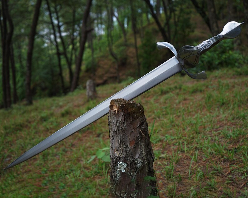 May include: A silver-toned metal sword with a double-edged blade, a crossguard with curved quillons, and a textured handle featuring a rounded pommel. The sword is balanced horizontally across the top of a rough, vertical wooden tree stump.