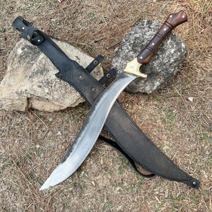 May include: A curved steel blade knife featuring a dark brown wooden handle with silver rivets and a brass bolster. The knife is accompanied by a black leather sheath with metal snap buttons and an adjustable strap for secure storage and transport.