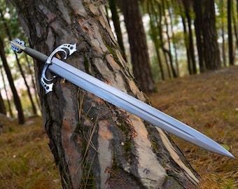 May include: A silver-toned metal sword featuring a double-edged blade, a textured grey grip, and an ornate, intricate crossguard with cut-out details. The pommel at the end of the handle matches the decorative style of the crossguard.