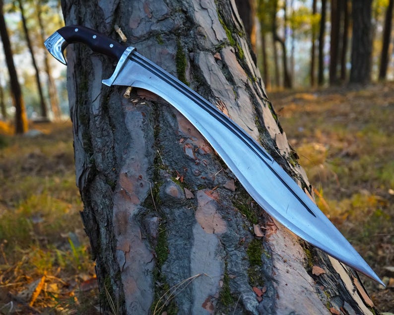 May include: A silver-colored sword with a black wooden handle and a curved blade. The sword is leaning against a tree trunk in a forest setting.
