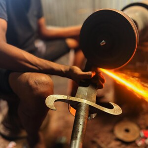 May include: A metal sword being sharpened against a rotating grinding wheel, producing a bright orange stream of sparks. The sword features a textured handle and a curved crossguard, while the grinding wheel is dark and circular in shape.