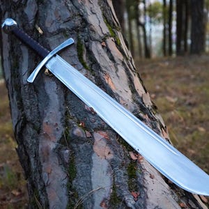 Puede incluir: Una espada de acero de doble filo con una empuñadura oscura y texturizada, un pomo redondeado y una guarda recta. La hoja es ancha con un canal central a lo largo, estrechándose hacia la punta y reflejando la luz en su superficie metálica.