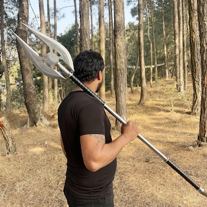 May include: A long polearm featuring a large, silver-colored metal head with three distinct, sharp-pointed blades. The shaft is constructed from a smooth, metallic material and includes two sections wrapped in black textured grip material for handling.