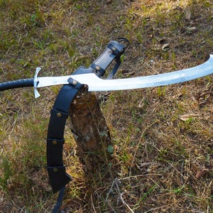 Puede incluir: Una espada plateada con una vaina de cuero negro y una hoja curva. La espada est&aacute; apoyada en un toc&oacute;n de &aacute;rbol en un &aacute;rea cubierta de hierba.
