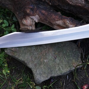 May include: A silver knife with a long, curved blade and a wooden handle. The knife is lying on a rock and surrounded by green grass.