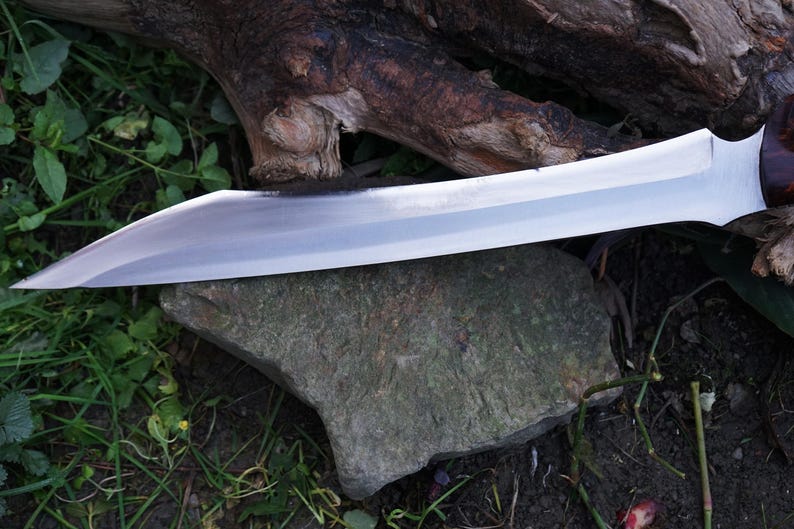 May include: A silver knife with a long, curved blade and a wooden handle. The knife is lying on a rock and surrounded by green grass.