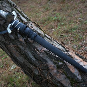 May include: A silver-toned sword with a dark, textured handle and a crossguard, encased in a black leather scabbard. The scabbard is secured with black leather straps and buckles, featuring ornate silver-colored metal fittings at the top and tip.
