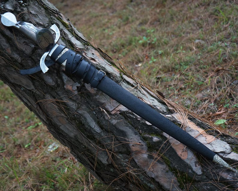 May include: A silver-toned sword with a dark, textured handle and a crossguard, encased in a black leather scabbard. The scabbard is secured with black leather straps and buckles, featuring ornate silver-colored metal fittings at the top and tip.