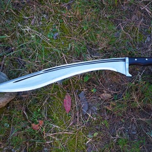May include: A silver-colored curved sword with a black wooden handle. The sword is lying on a bed of green grass and brown leaves.