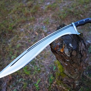 May include: A silver-colored sword with a black handle and a curved blade. The sword is resting on a tree stump.