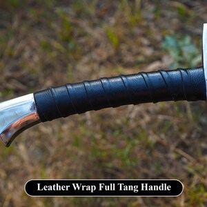 Puede incluir: Primer plano de un mango de espada plateado envuelto en cuero negro. El mango es curvo y tiene un acabado liso y pulido. El texto "Leather Wrap Full Tang Handle" es visible en la parte inferior de la imagen.