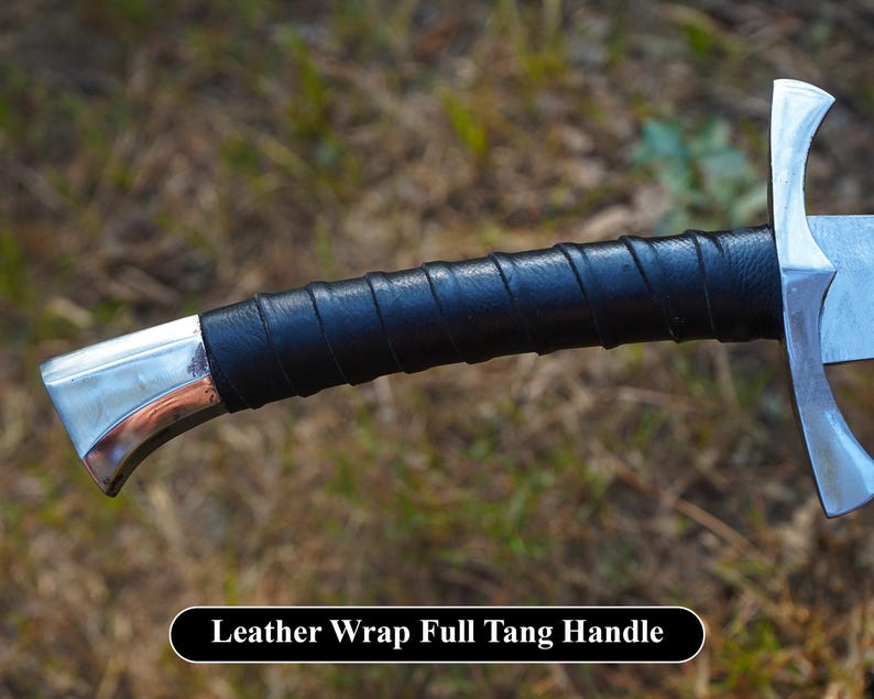Puede incluir: Primer plano de un mango de espada plateado envuelto en cuero negro. El mango es curvo y tiene un acabado liso y pulido. El texto "Leather Wrap Full Tang Handle" es visible en la parte inferior de la imagen.