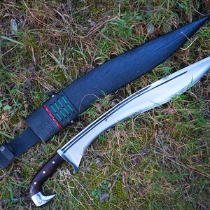 May include: A silver blade with a brown wooden handle and a black leather sheath with green and red stitching. The blade is lying on a bed of green grass.