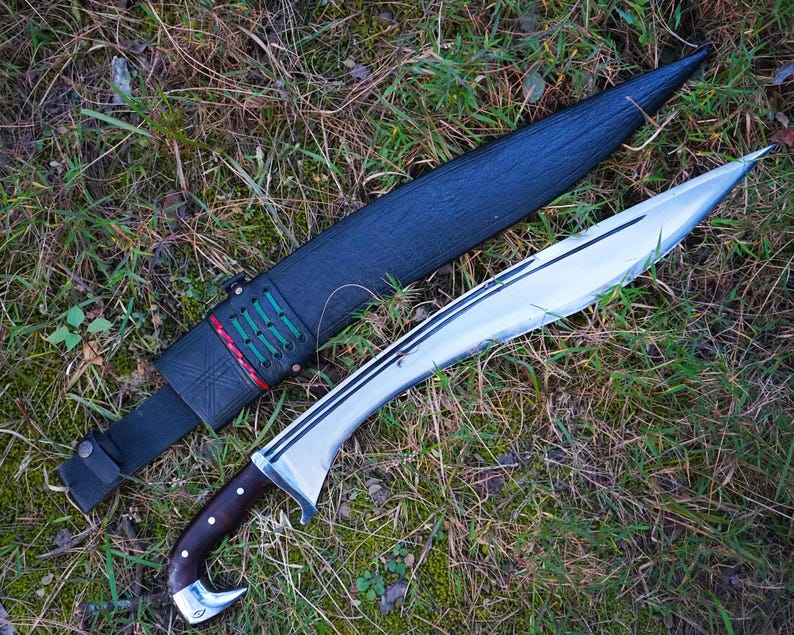 May include: A silver blade with a brown wooden handle and a black leather sheath with green and red stitching. The blade is lying on a bed of green grass.