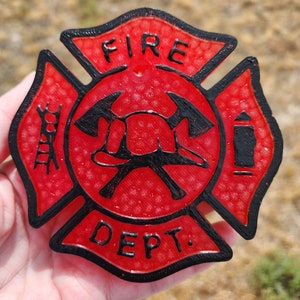 May include: A red and black Maltese cross emblem featuring the text FIRE DEPT. The center displays a firefighter helmet and two crossed axes. The left side shows a ladder icon, and the right side shows a fire hydrant icon, all with a textured finish.