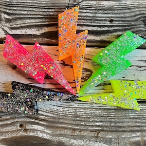 May include: Five glitter lightning bolt ornaments in black, pink, green, yellow, and orange. The ornaments are hanging from a wooden surface.