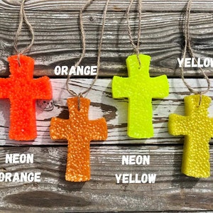 May include: Four hanging cross-shaped air fresheners in neon orange, neon yellow, orange, and yellow. The crosses are made of a textured material and have a string for hanging.