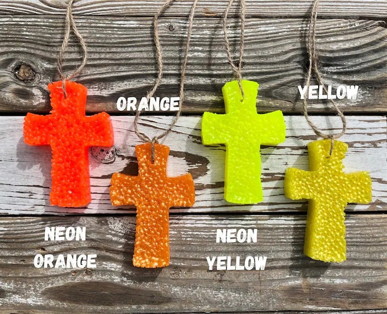 May include: Four hanging cross-shaped air fresheners in neon orange, neon yellow, orange, and yellow. The crosses are made of a textured material and have a string for hanging.