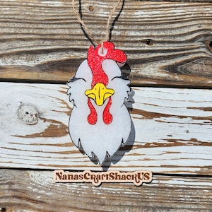 May include: A white chicken head ornament with a red glittery comb and wattle, a yellow beak, and black eye details. The ornament hangs from a jute twine string and features a wooden sign below it with the text NanasCraftShackUS printed in black.
