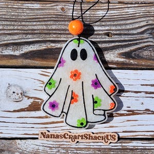 May include: A white ghost-shaped air freshener with black eyes and colorful floral accents in orange, purple, and green. It hangs from a black string with an orange bead. The background is a rustic wooden surface. The text "NanasCraftShackUS" is at the bottom.