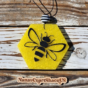 May include: A yellow hexagon-shaped pendant with a black silhouette of a bee. The pendant is hanging from a black cord with a black and white striped bead.