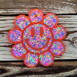 May include: A bright orange, flower-shaped decoration with a smiling face. The petals and face are filled with colorful glitter. The decoration is set against a weathered, wooden background.