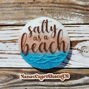 May include: Round coaster with the phrase "salty as a beach" in brown script. The coaster features a gradient design, transitioning from white to tan, with a blue wave pattern at the bottom. The shop name "NanasCraftShackUS" is at the bottom.