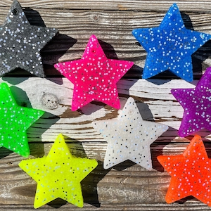 May include: A collection of star-shaped air fresheners in various colors, including black, pink, blue, green, yellow, white, purple, and orange. Each star is covered in glitter, creating a sparkling effect. The air fresheners are displayed on a wooden surface.