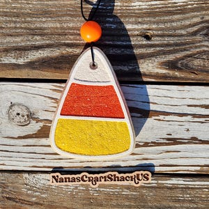 May include: A triangular candy corn ornament featuring horizontal bands of white, orange, and yellow textured material. A black cord with a single bright orange bead is attached to the top. A wooden sign below reads NanasCraftShackUS.