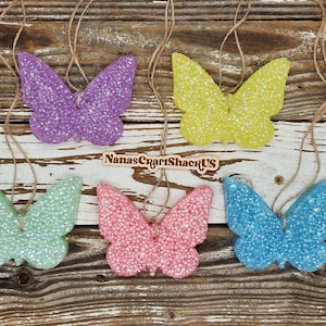 May include: Five butterfly-shaped ornaments with a glittery texture in purple, yellow, light green, pink, and blue. Each ornament hangs from a brown twine string. A small wooden sign with the text NanasCraftShackUS is positioned in the center.