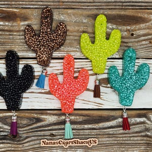 May include: Five colorful cactus-shaped air fresheners with tassels hanging from them. The colors are black, brown, green, orange, and blue. The background is a wooden surface.