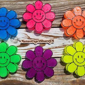 May include: Six colorful resin flower-shaped coasters with a black outline and a smiley face in the center. The coasters are arranged in a circle on a wooden surface. The text "NanasCraftShackUS" is visible in the center of the image.