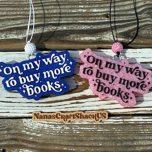 May include: Two glittery hanging ornaments with the text On my way to buy more books. One is dark blue with white text and a white bead, the other is light pink with black text and a pink bead. A wooden sign reads NanasCraftShackUS below them.