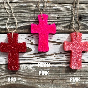 May include: Three cross-shaped car air fresheners hanging from twine. The crosses are made of a textured material and are in red, pink, and neon pink.