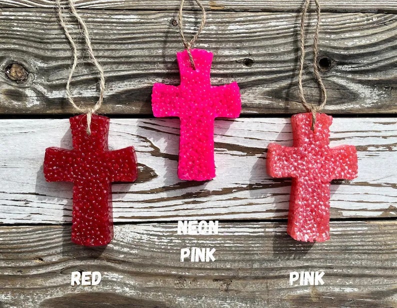 May include: Three cross-shaped car air fresheners hanging from twine. The crosses are made of a textured material and are in red, pink, and neon pink.