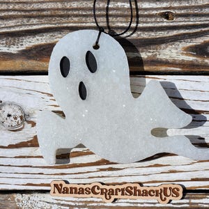 May include: A white glittery ghost-shaped ornament with two oval eyes and an oval mouth in black. The ornament features a black string loop at the top for hanging. Below the ghost, a wooden sign displays the text NanasCrartShackUS.