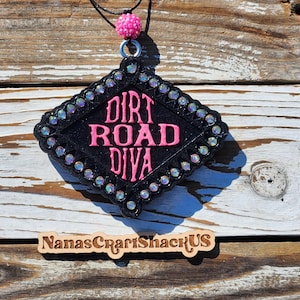 May include: Black, diamond-shaped pendant with the text "DIRT ROAD DIVA" in pink. The pendant is framed with iridescent rhinestones and a beaded pink accent. A black cord and pink bead are attached for hanging.
