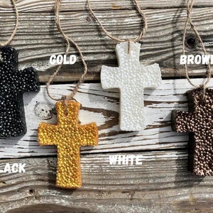 May include: Four decorative crosses made of a textured material in black, white, gold, and brown. The crosses are hanging from twine and are displayed against a wooden background. The words "BLACK", "GOLD", "WHITE", and "BROWN" are written below each cross.