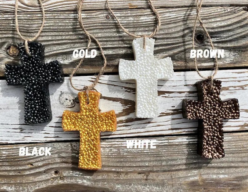 May include: Four decorative crosses made of a textured material in black, white, gold, and brown. The crosses are hanging from twine and are displayed against a wooden background. The words "BLACK", "GOLD", "WHITE", and "BROWN" are written below each cross.