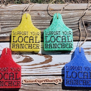 May include: Four colorful, textured cow tag ornaments. Each tag is a different color: yellow, green, red, and blue. The black text on each tag reads "SUPPORT YOUR LOCAL RANCHER". Each tag has a twine loop for hanging.