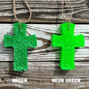 May include: Two green cross-shaped car air fresheners hanging from twine. The cross on the left is a solid green color, while the cross on the right is a neon green color. The text "GREEN" is below the left cross and "NEON GREEN" is below the right cross.