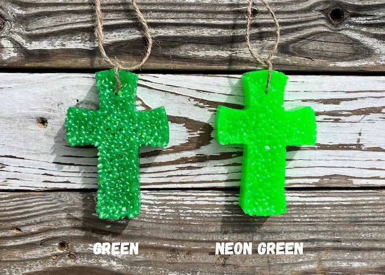 May include: Two green cross-shaped car air fresheners hanging from twine. The cross on the left is a solid green color, while the cross on the right is a neon green color. The text "GREEN" is below the left cross and "NEON GREEN" is below the right cross.