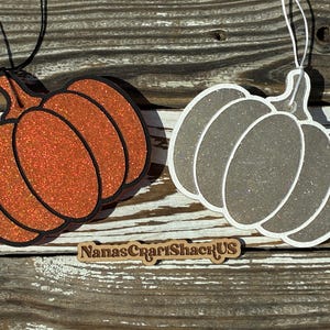 May include: Two pumpkin-shaped hanging ornaments, one orange with black outlines and one grey with white outlines, both featuring a glittery finish. Each ornament has a string attached at the top. A wooden sign with the text NanasCraftShackUS is present.